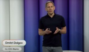 Dmitri Dolgov, co-CEO da Waymo, fala em um evento sobre tecnologia, vestindo camiseta preta e jeans, em frente a um fundo com luzes azuladas e roxas.