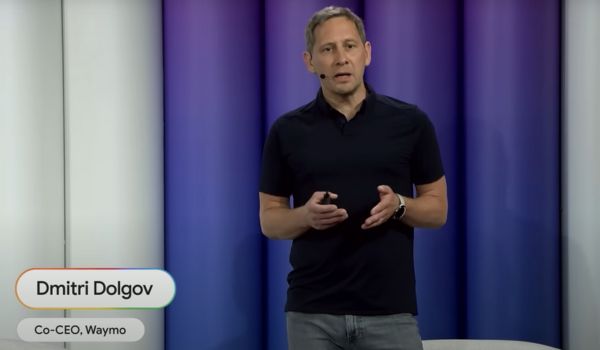Dmitri Dolgov, co-CEO da Waymo, fala em um evento sobre tecnologia, vestindo camiseta preta e jeans, em frente a um fundo com luzes azuladas e roxas.