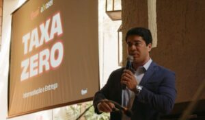 Design seHomem em traje social fala ao microfone durante apresentação, ao lado de uma tela com os dizeres "TAXA ZERO – Intermediação e Entrega".m nome