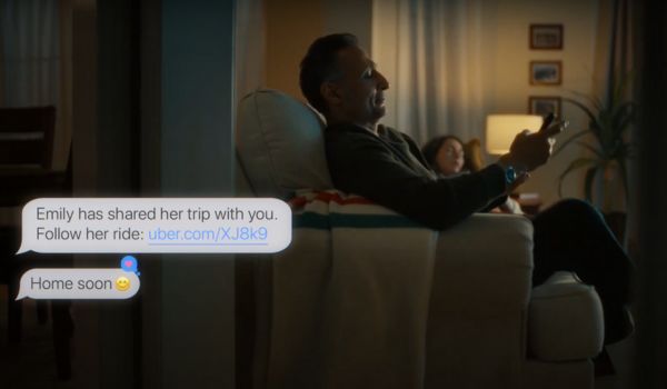 Homem de meia-idade sentado em uma poltrona usando o celular, com uma mulher deitada no sofá ao fundo. Na imagem, aparecem duas mensagens sobrepostas: a primeira informa que "Emily compartilhou a viagem com você" e fornece um link para acompanhar a corrida; a segunda diz "Chego logo ?".