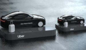 Dois troféus em formato de carros pretos futuristas com a inscrição "Uber Airport Awards", cada um acompanhado por uma mala cinza em miniatura, posicionados sobre bases com o logo da Uber.