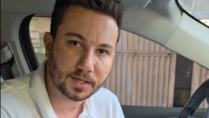 Homem de barba curta, vestindo camisa polo branca com logotipo “GoHop”, sentado no banco do motorista de um carro moderno, olhando diretamente para a câmera enquanto fala. Ele segura as mãos em frente ao corpo e um celular com texto visível está apoiado sobre a perna.