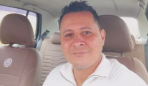 Homem sorrindo, usando uma blusa branca, sentado no banco do motorista de um carro com interior marrom da Volkswagen.