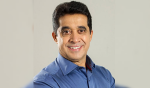 Retrato de um homem sorrindo, com cabelo curto e escuro, usando uma camisa azul clara. Ele está olhando para a câmera com um sorriso amigável, destacando uma expressão confiável e positiva.