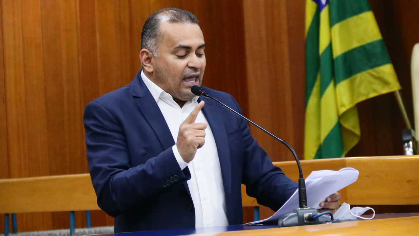 Homem de terno azul-marinho e camisa branca fala ao microfone em uma tribuna de madeira, segurando papéis na mão direita e gesticulando com a esquerda, com bandeira do estado de Goiás ao fundo.