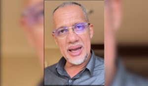 Homem de pele parda, com cabelo grisalho curto e barba branca rala, usando óculos com lentes azuladas e camisa social escura, falando diretamente para a câmera em ambiente interno.