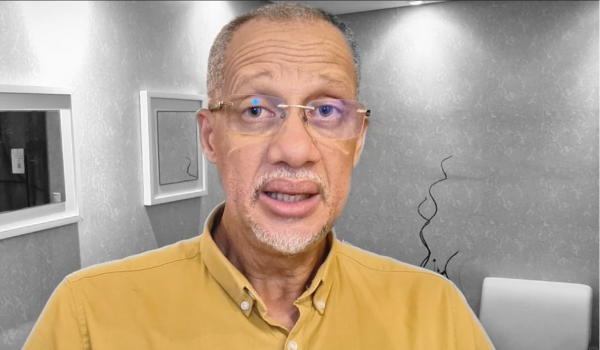 Homem de pele clara, cabelos curtos e barba, vestindo uma camisa amarela, com óculos, em um ambiente com paredes decoradas com papel de parede cinza e quadros brancos.