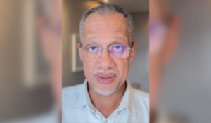 Retrato de um homem de pele clara, com cabelo curto e grisalho, usando óculos de armação fina e uma camisa branca. Ele olha diretamente para a câmera, com uma expressão séria.