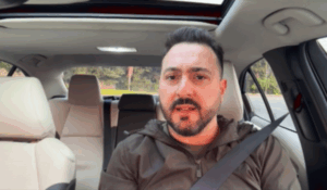 Homem de barba curta e cabelo escuro está sentado no banco do motorista de um carro moderno, usando cinto de segurança. Ele veste uma jaqueta esportiva marrom e parece estar falando, possivelmente gravando um vídeo.