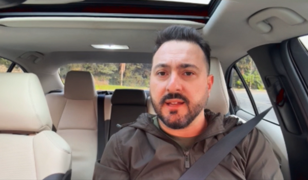 Homem de barba curta e cabelo escuro está sentado no banco do motorista de um carro moderno, usando cinto de segurança. Ele veste uma jaqueta esportiva marrom e parece estar falando, possivelmente gravando um vídeo.