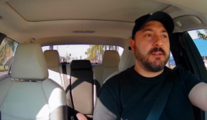 Homem dirigindo um carro, com uma expressão séria, usando uma camiseta preta e boné, em um ambiente claro e ensolarado. O banco do passageiro está vazio, e a cidade é visível através das janelas.