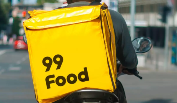 Entregador de moto com mochila amarela da 99 Food em uma rua urbana durante o dia.