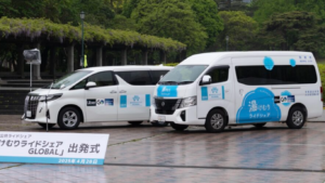 Dois veículos brancos com adesivos da Uber e do projeto "Yukemuri Ride Share GLOBAL" estão estacionados em uma praça molhada pela chuva, em frente a uma placa com a data de lançamento do serviço: 28 de abril de 2025.