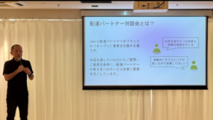 Homem com microfone se apresenta diante de uma cortina branca ao lado de um telão que exibe informações em japonês sobre uma reunião entre a Uber e parceiros de entrega.