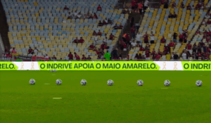 Placar eletrônico de estádio de futebol exibindo a mensagem "O inDrive apoia o Maio Amarelo", com bolas de futebol alinhadas no gramado e arquibancadas parcialmente ocupadas ao fundo.