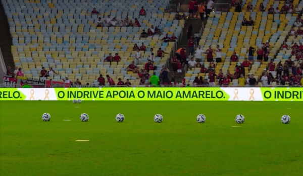 Placar eletrônico de estádio de futebol exibindo a mensagem "O inDrive apoia o Maio Amarelo", com bolas de futebol alinhadas no gramado e arquibancadas parcialmente ocupadas ao fundo.