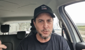 Homem de barba e boné preto com a inscrição “NYC” fala sério dentro de um carro.