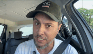 Homem de boné preto com logo "NYC", barba por fazer e camisa branca, falando diretamente para a câmera de dentro de um carro.