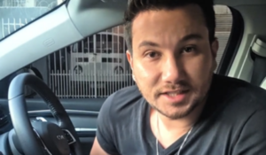 Homem jovem com barba rala e cabelo curto, usando camiseta preta, sentado no banco do motorista de um carro e olhando diretamente para a câmera.