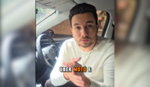 Homem com cabelo curto e barba, usando uma camisa branca, sentado no banco do motorista de um carro. Ele está fazendo um gesto com as mãos enquanto olha para a câmera. A palavra "UBER MOTO E" está visível na parte inferior da imagem.