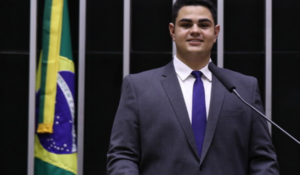 Homem jovem de terno fala em um púlpito com microfone, ao lado da bandeira do Brasil.