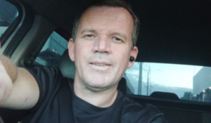 Homem branco de cabelo curto e grisalho tirando uma selfie dentro do carro, usando camiseta preta e fone de ouvido no ouvido direito. Ele está com cinto de segurança e sorri levemente para a câmera.