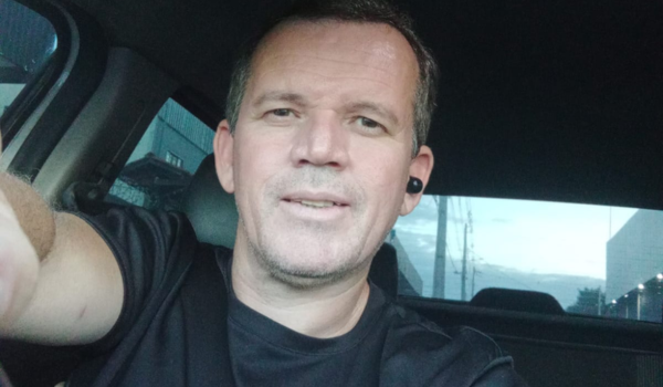 Homem branco de cabelo curto e grisalho tirando uma selfie dentro do carro, usando camiseta preta e fone de ouvido no ouvido direito. Ele está com cinto de segurança e sorri levemente para a câmera.