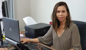 Mulher sentada à mesa de escritório, sorrindo para a câmera, com notebook, teclado e impressora ao fundo.