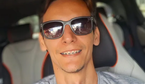 Homem com óculos escuros sorrindo dentro de um carro, vestindo uma camiseta cinza.