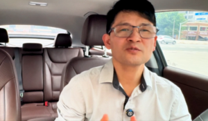 Homem de pele clara, cabelo preto e óculos, usando camisa social branca, falando enquanto está sentado no banco da frente de um carro; o interior do veículo é marrom e a rua está visível pela janela.