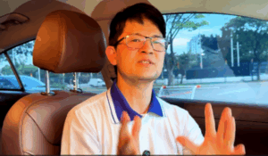 Homem de óculos, vestindo uma camisa polo branca com detalhes azuis, fala com expressividade enquanto está sentado no banco do motorista de um carro com interior marrom.