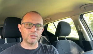 Homem de óculos e camiseta escura sentado no banco do motorista de um carro, com expressão séria, olhando para a câmera durante o dia.