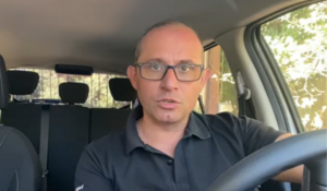 Motorista de aplicativo Marcelo Ribeiro falando de dentro de seu carro, usando camisa preta e óculos.