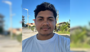 Homem de pele morena, cabelo preto cacheado e barba rala, usando camiseta branca, tira uma selfie ao ar livre em um dia ensolarado. Ao fundo, há céu azul, coqueiros, calçada, postes e algumas casas.