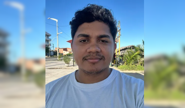 Homem de pele morena, cabelo preto cacheado e barba rala, usando camiseta branca, tira uma selfie ao ar livre em um dia ensolarado. Ao fundo, há céu azul, coqueiros, calçada, postes e algumas casas.