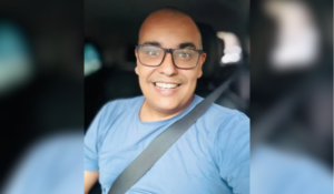 Homem sorridente de óculos e camiseta azul tira uma selfie dentro de um carro, usando cinto de segurança.