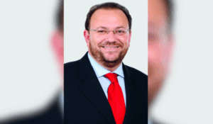 Homem de meia-idade sorridente, com barba e óculos, vestindo terno preto, camisa azul clara e gravata vermelha, posando para uma foto em fundo branco.