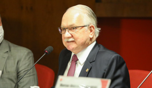 Homem idoso de cabelo grisalho e penteado para trás, usando terno escuro, camisa branca e gravata vermelha, fala ao microfone em uma mesa de reunião. Ele usa óculos e está em ambiente interno com iluminação quente. À esquerda, outra pessoa parcialmente visível usa máscara.