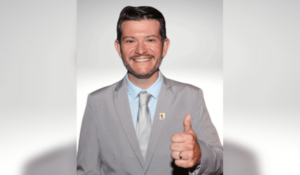 Homem sorrindo, usando terno cinza claro, com camisa e gravata prata, fazendo um sinal de positivo com a mão direita. Ele está em um fundo neutro.