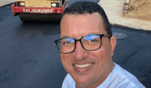 Homem sorridente usando óculos e camiseta branca tira uma selfie em uma rua recém-asfaltada, com um rolo compressor ao fundo.