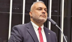 Homem de terno escuro e gravata vermelha fala em um microfone em ambiente institucional com barras verticais ao fundo.