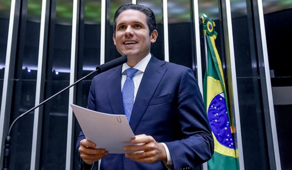 Homem de terno azul escuro fala ao microfone em um púlpito com a bandeira do Brasil ao fundo.