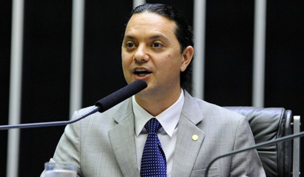 Um homem de terno cinza claro fala ao microfone em uma sessão na Câmara dos Deputados. Ele está com um olhar atento e usa uma gravata de pontos brancos.