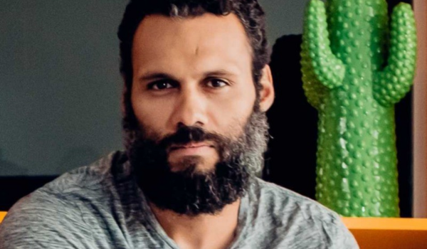 Homem de barba cheia e cabelo curto, vestindo camiseta cinza, sentado próximo a uma decoração de cacto verde.