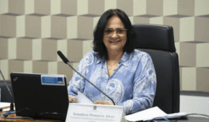 Senadora Damares Alves sentada à mesa durante sessão no Senado Federal, sorrindo, com papéis à frente e um notebook com o selo do Senado.