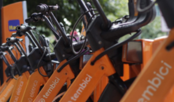 Bicicletas elétricas compartilhadas da Tembici estacionadas em fila, com detalhes em laranja e preto, prontas para uso em estação próxima a áreas verdes.