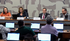 Quatro pessoas sentadas atrás de uma bancada durante uma audiência pública no Senado. À frente, várias pessoas com laptops acompanham a sessão. Ao fundo, uma parede com padrão geométrico.