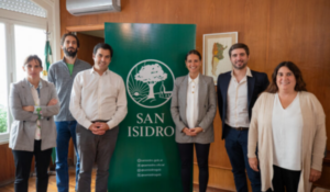 Grupo de sete pessoas posa sorrindo ao lado de um banner verde com o logotipo e o nome "San Isidro", em uma sala com parede de madeira.