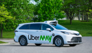 Carro branco autônomo com logotipo “UberMAY” estacionado em via asfaltada, cercado por árvores verdes em um parque.