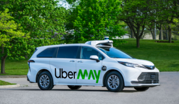Carro branco autônomo com logotipo “UberMAY” estacionado em via asfaltada, cercado por árvores verdes em um parque.